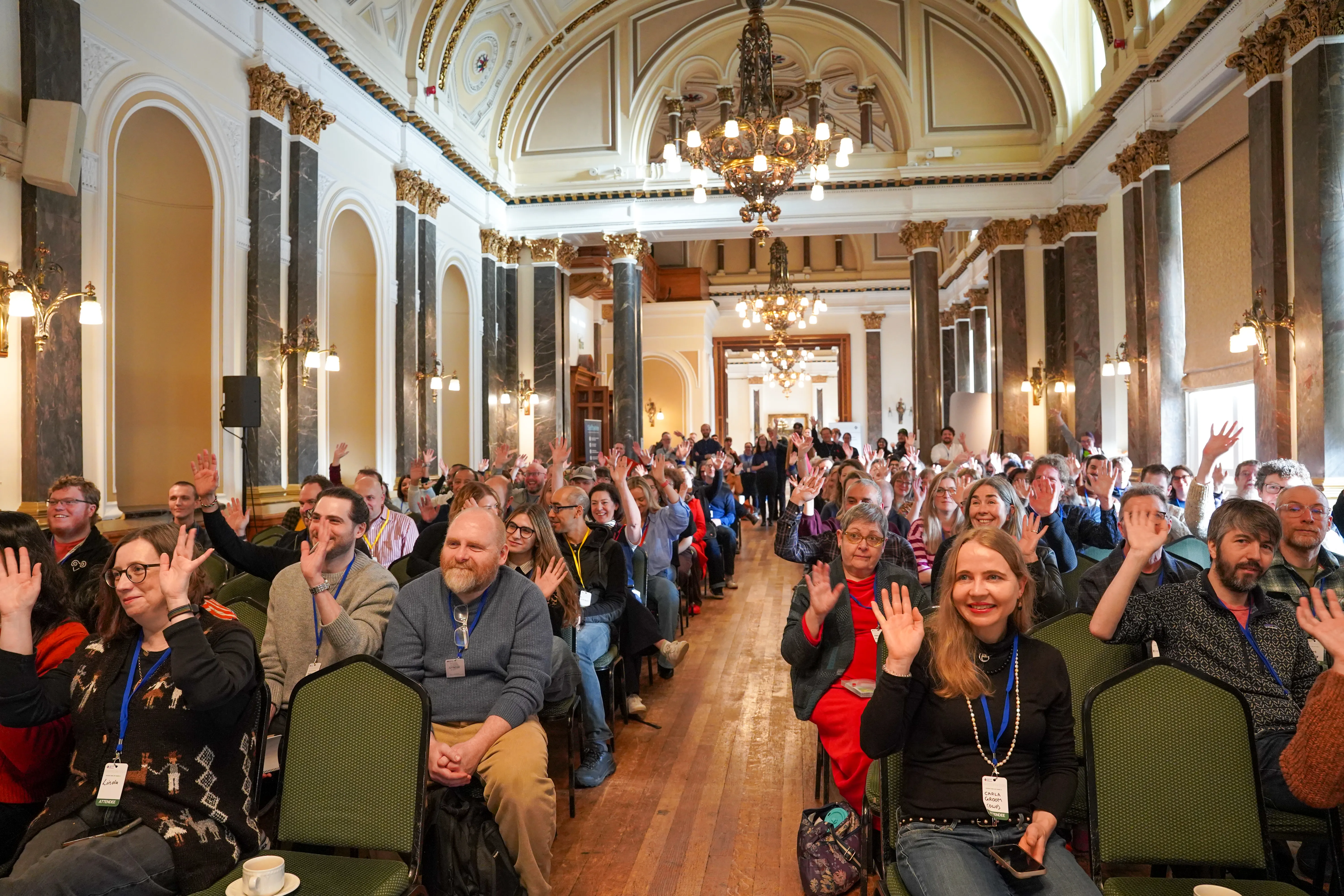 Wide shot of a crowded hall where approximately 100 people are seated. Many participants have their hands raised toward the stage, signaling their interest in pitching. The atmosphere is professional and collaborative.